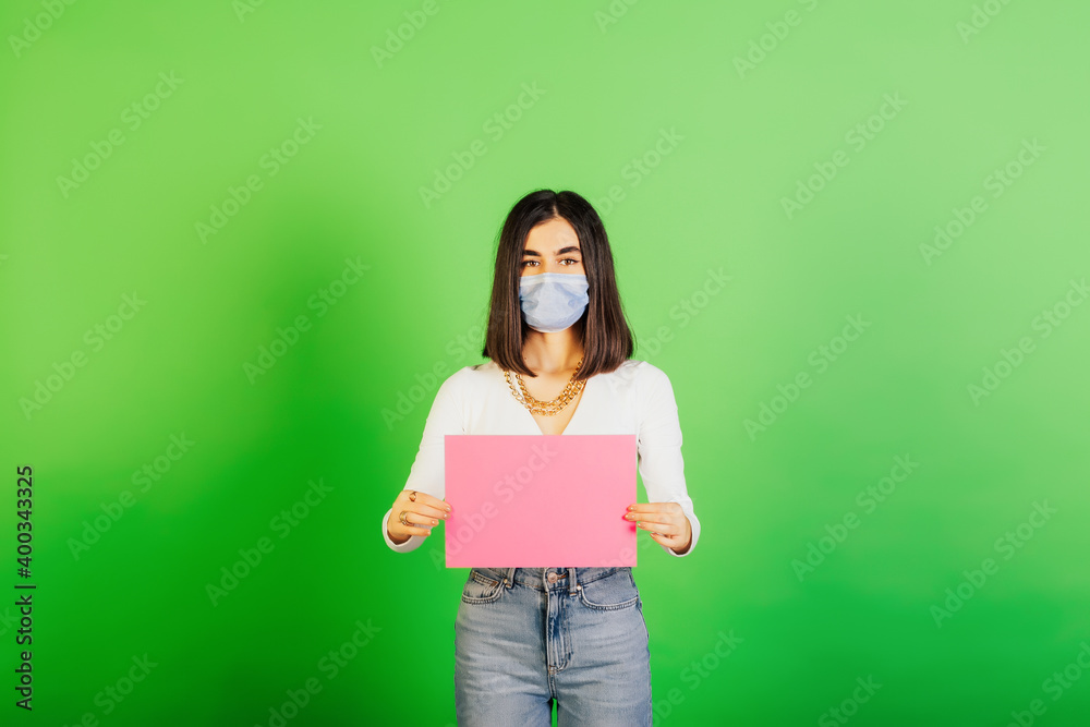 Woman in medical mask with pink blank sheet of paper isolated on green background. Stay at home, quarantine. Copy space. Woman in medical mask with pink blank sheet of paper isolated on green background. Stay at home, quarantine. Copy space.
