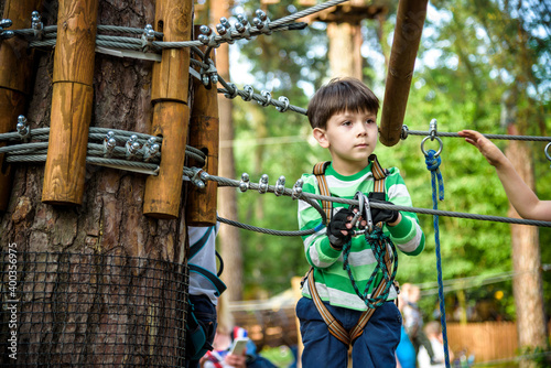 Young cute child boy in summer clothing, safety harness and helmet attached with carbine to cable moves slowly along rope way on green trees sunny bokeh background. Sport, game, leisure concept