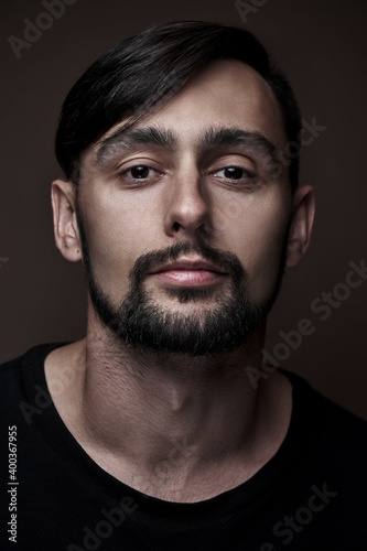 Portrait of handsome caucasian man with beard and stylish haircut in black clothes posing on brown background isolated in studio, fashion model looking at the camera