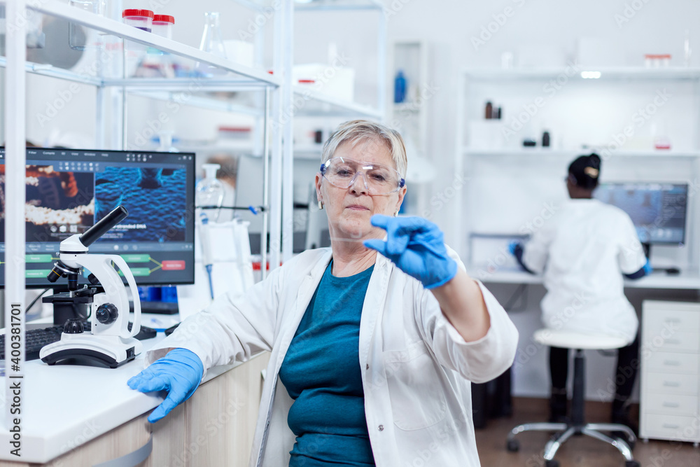 Senior scientist in modern laboratory holding genetic sample on laboratory glass. Elderly researcher in sterile lab looking on microscope slide wearing lab coat.