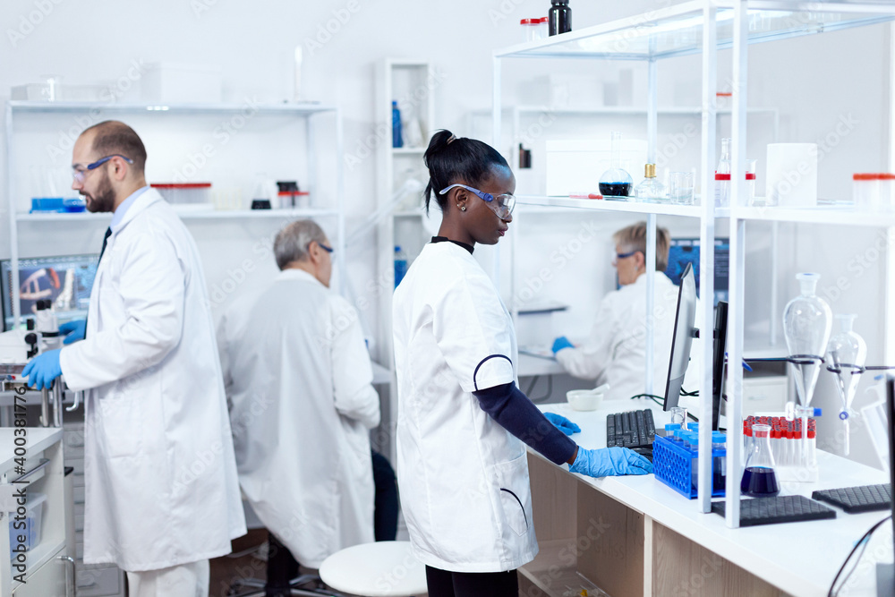 African scientist and her colleagues doing experiments in sterile laboratory. Multiethnic team of researchers working in microbiology lab testing solution for medical purpose. African scientist and her colleagues doing experiments in sterile laboratory. Multiethnic team of researchers working in microbiology lab testing solution for medical purpose.