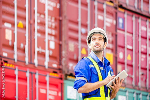 Engineer or Technician wears white hardhat and reflection shirt and holding tablet and look at sky and smile with cargo stacked containers in background. Concept of inventory and logistic management.
