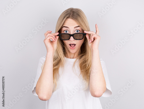 Portrait of a surprised young beautiful girl in eyeglasses looking at camera while raised up glasses. Isolated over gray background