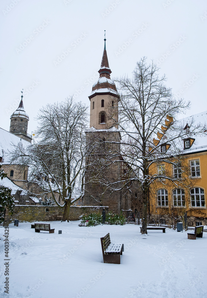 Snow covered park with winter trees & an old church spire with traditional painted building in yellow. Taken in Feuchtwangen, Germany in winter