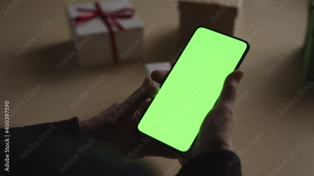 Handheld shot of old female hands holding smartphone with green screen over wood vintage table with presents on background and dim window light Handheld shot of old female hands holding smartphone with green screen over wood vintage table with presents on background and dim window light