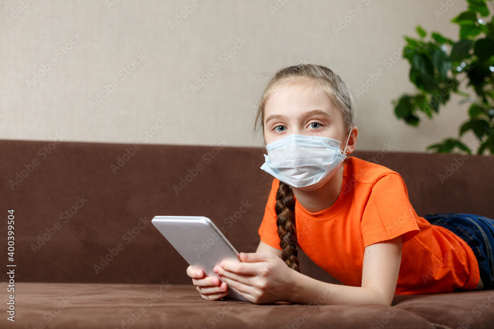 Little girl in medical mask lying on the sofa and using tablet pc. Isolation at home. Looking into the camera.