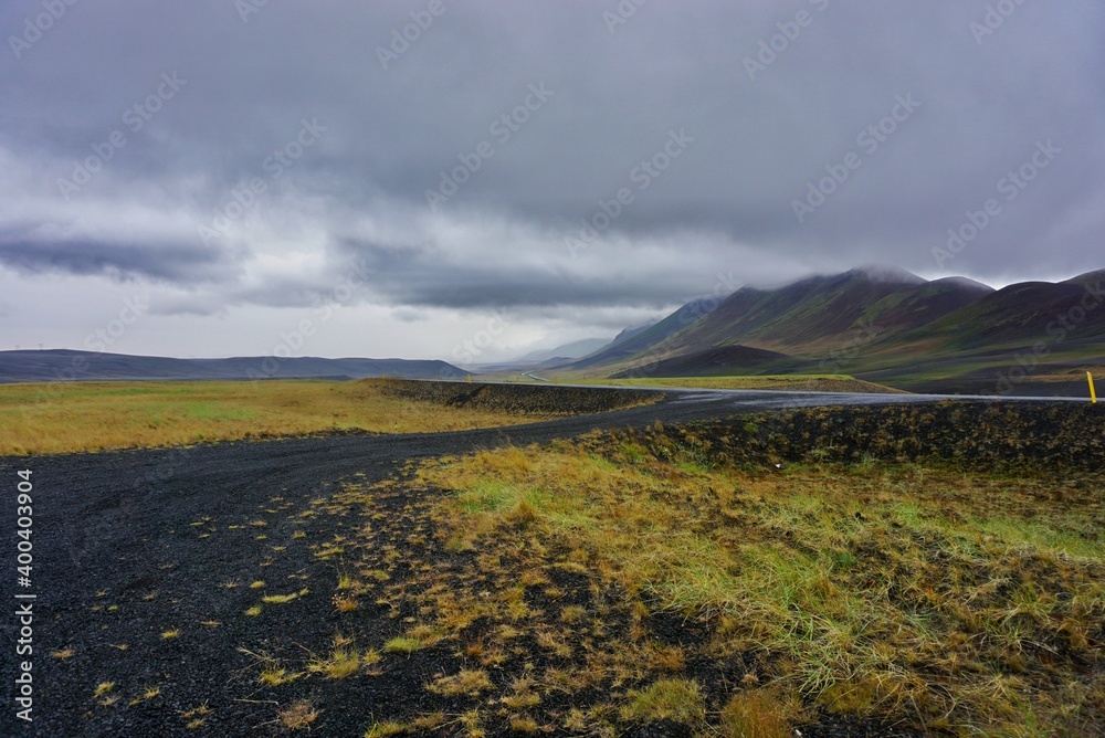 yellow grass grows on a black volcanic field, colorful mountains are in the background, heavy clouds touch the top of the mountains, nature of Iceland