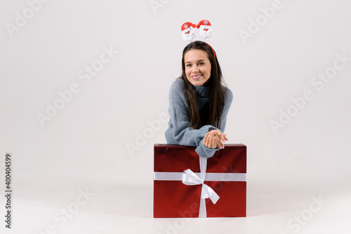 Happy excited woman in funny head hoop staying near the red big gift christmas box isolated on the gray background