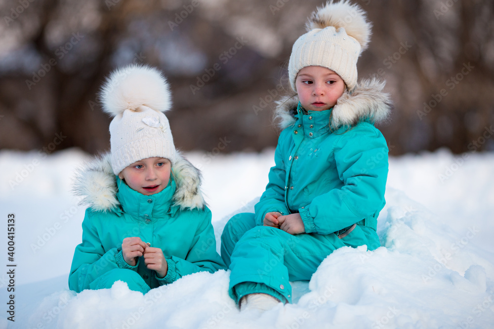 Two funny little girls in identical clothes are having fun in a beautiful winter park. Cute kids playing in the snow. Winter activities for children.