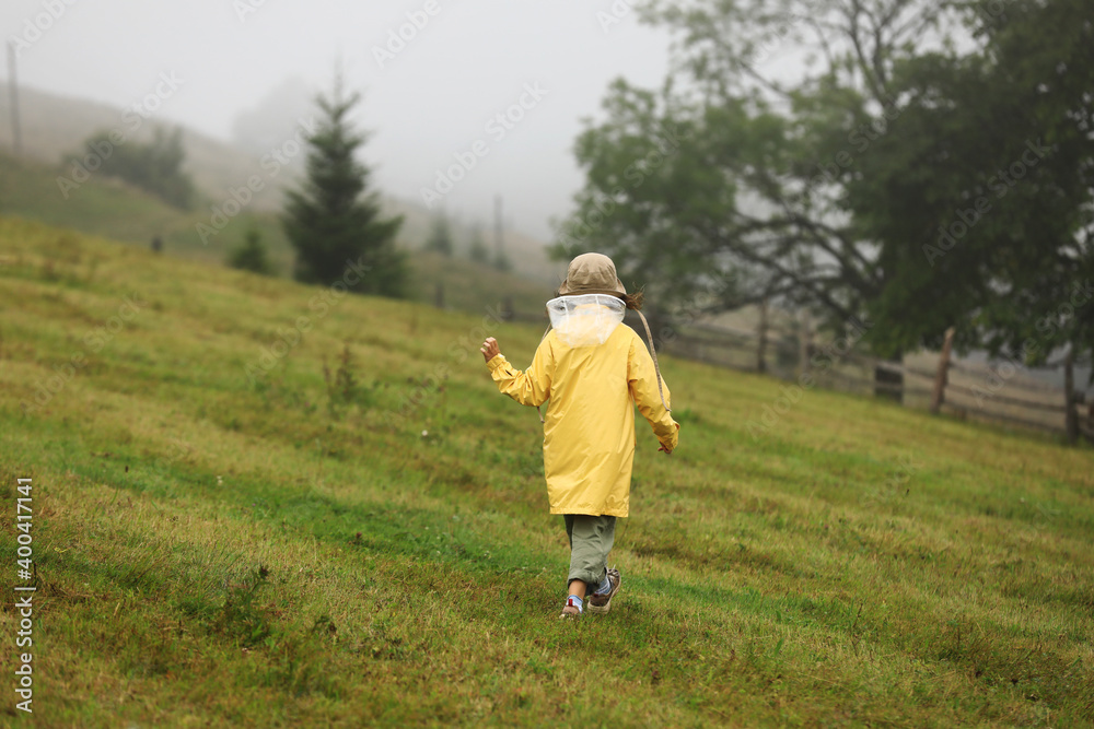Cute little girl in yellow raincoat is walking in foggy mountains. Bad weather, summer tropical storm, autumn fashion concept. back view
