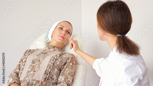 Young woman in white coat talks to mature lady showing places of cosmetic operation sitting in white chair in corner of beauty salon ward