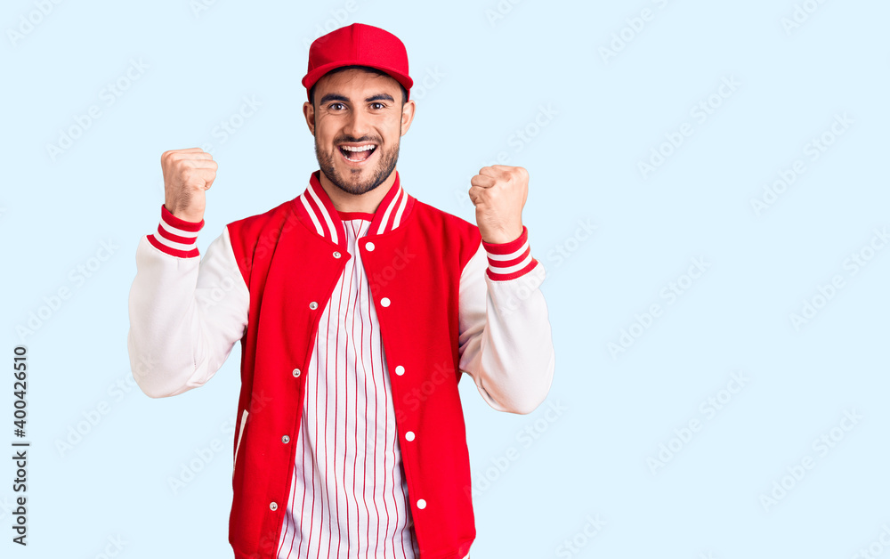 Young handsome man wearing baseball jacket and cap screaming proud, celebrating victory and success very excited with raised arms Young handsome man wearing baseball jacket and cap screaming proud, celebrating victory and success very excited with raised arms