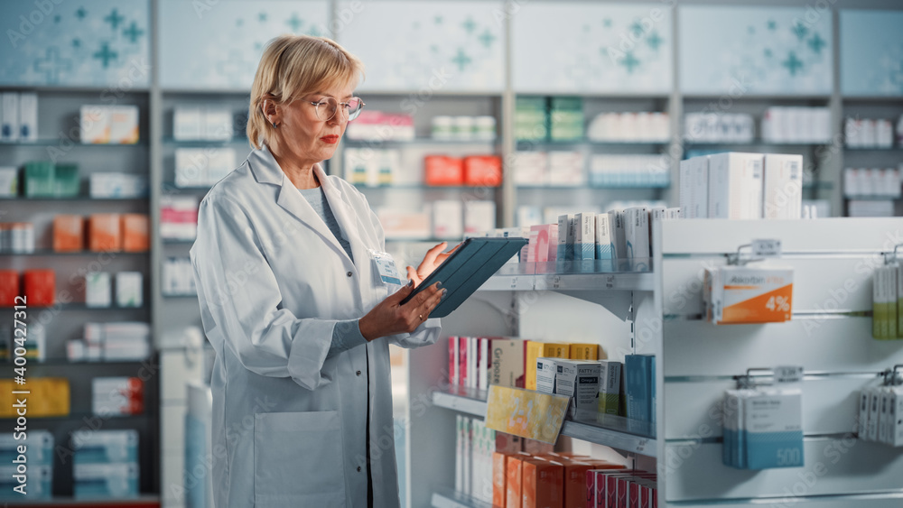 Pharmacy Drugstore: Mature Experienced Female Farmacist Uses Digital Tablet Computer to Arrange, Inventory, Check Medicine, Drugs, Vitamins on a Shelf. Medical Professional in Pharma Shop