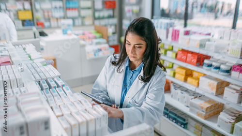 Pharmacy Drugstore: Beautiful Latina Pharmacist Uses Digital Tablet Computer, Checks Inventory of Medicine, Drugs, Vitamins, Health Care Products on a Shelf. Professional Pharmacist in Pharma Store