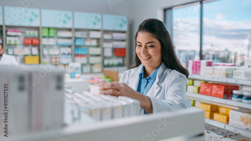 фотографія Pharmacy Drugstore: Portrait of Beautiful Latina Pharmacist Wearing White Coat Arranges Medicine, Drugs, Vitamins on a Shelf