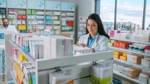 Pharmacy Drugstore: Portrait of Beautiful Latina Pharmacist Wearing White Coat Arranges Medicine, Drugs, Vitamins on a Shelf. Professional Pharmacist in Pharma Store Shelves with Health Care Products