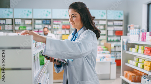 Pharmacy Drugstore  Beautiful Latina Pharmacist Uses Digital Tablet Computer  Checks Inventory of Medicine  Drugs  Vitamins  Health Care Products on a Shelf. Professional Pharmacist in Pharma Store