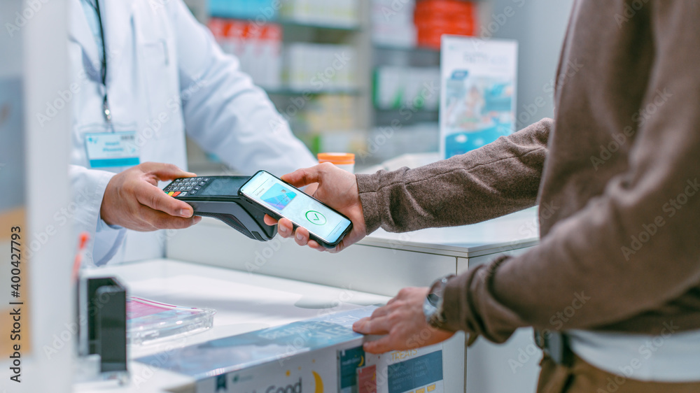 Pharmacy Drugstore Checkout Cashier Counter: Senior Latin Pharmacist and Male Customer Paying with NFC Contactless Smartphone for Prescription Medicine, Vitamins. Modern Store with Health Care Product