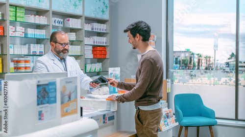 Fotografie Pharmacy Drugstore Checkout Cashier Counter: Senior Latin Pharmacist and Male Customer Paying with NFC Contactless Smartphone for Prescription Medicine, Vitamins