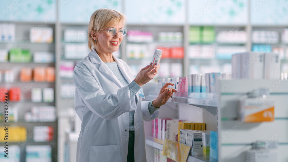Pharmacy Drugstore: Mature Experienced Female Farmacist Arranges Inventory, Check Medicine, Drugs, Vitamins on a Shelf. She Holds Box with Best Medicinal Pills. Medical Professional in Pharma Shop