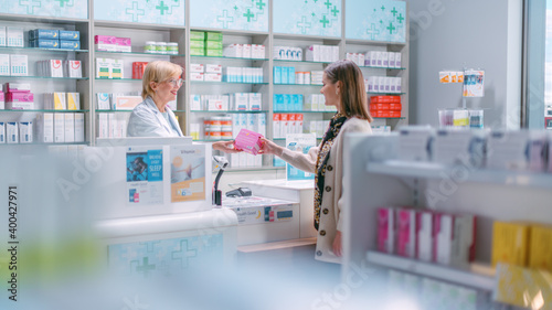 Pharmacy Drugstore Checkout Cashier Counter: Mature Female Pharmacist Passes Pink Box with Cure to a Female Customer, who is Buying Prescription Medicine, Vitamins, Beauty, Health Care Products.