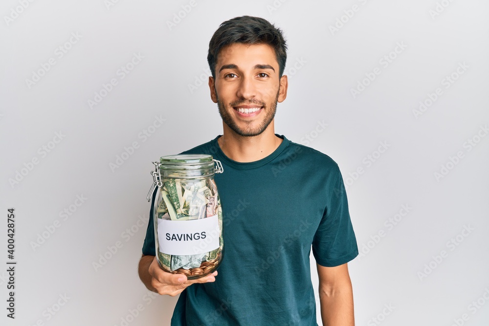 Young handsome man holding jar with savings looking positive and happy standing and smiling with a confident smile showing teeth Young handsome man holding jar with savings looking positive and happy standing and smiling with a confident smile showing teeth