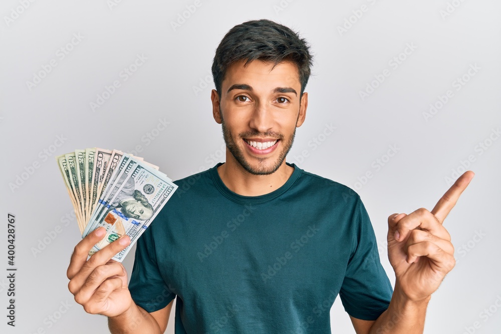 Young handsome man holding dollars smiling happy pointing with hand and finger to the side Young handsome man holding dollars smiling happy pointing with hand and finger to the side