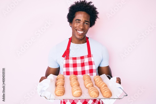 Handsome african american man with afro hair wearing baker uniform holding homemade bread winking looking at the camera with sexy expression, cheerful and happy face.