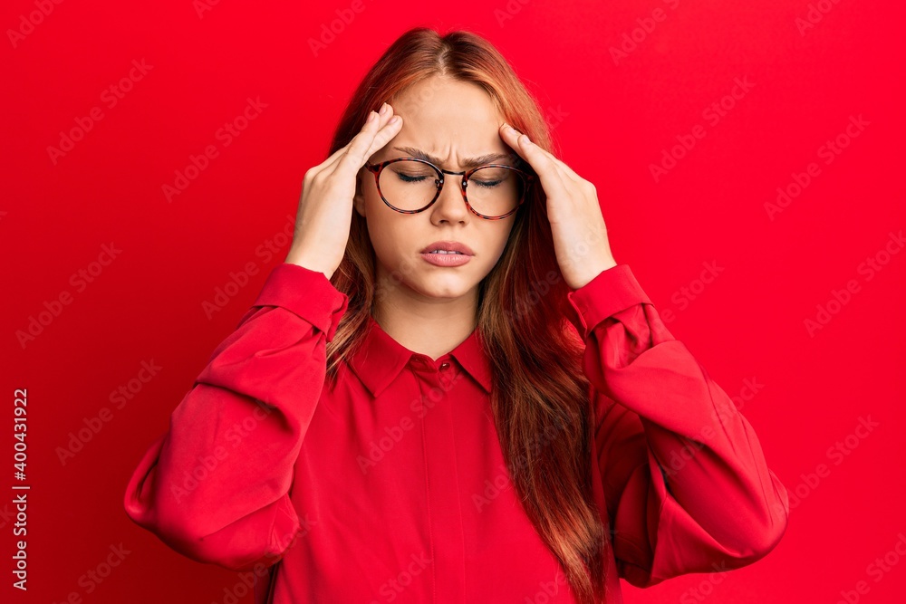 Young beautiful redhead woman wearing casual clothes and glasses over red background with hand on head for pain in head because stress. suffering migraine. Young beautiful redhead woman wearing casual clothes and glasses over red background with hand on head for pain in head because stress. suffering migraine.