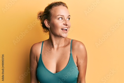 Beautiful caucasian woman wearing sleeveless shirt over yellow background looking away to side with smile on face, natural expression. laughing confident.