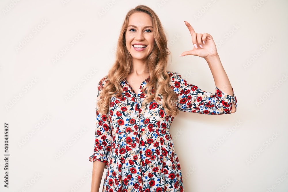 Young caucasian woman with blond hair wearing summer dress smiling and confident gesturing with hand doing small size sign with fingers looking and the camera. measure concept. Young caucasian woman with blond hair wearing summer dress smiling and confident gesturing with hand doing small size sign with fingers looking and the camera. measure concept.