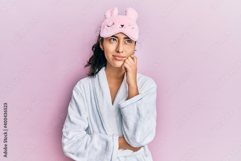 Hispanic teenager girl with dental braces wearing sleep mask and robe serious face thinking about question with hand on chin, thoughtful about confusing idea