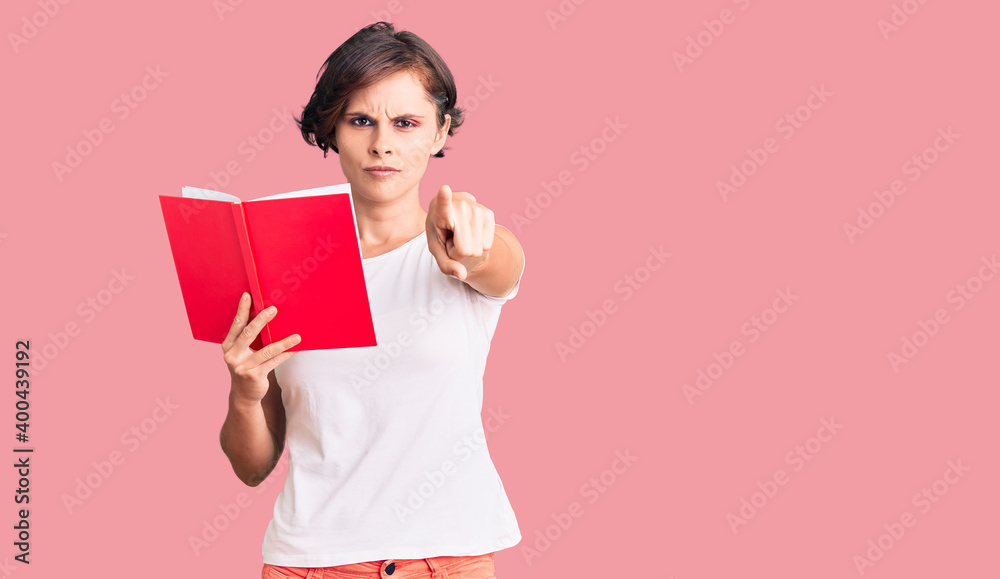 Beautiful young woman with short hair reading a book pointing with finger to the camera and to you, confident gesture looking serious Beautiful young woman with short hair reading a book pointing with finger to the camera and to you, confident gesture looking serious