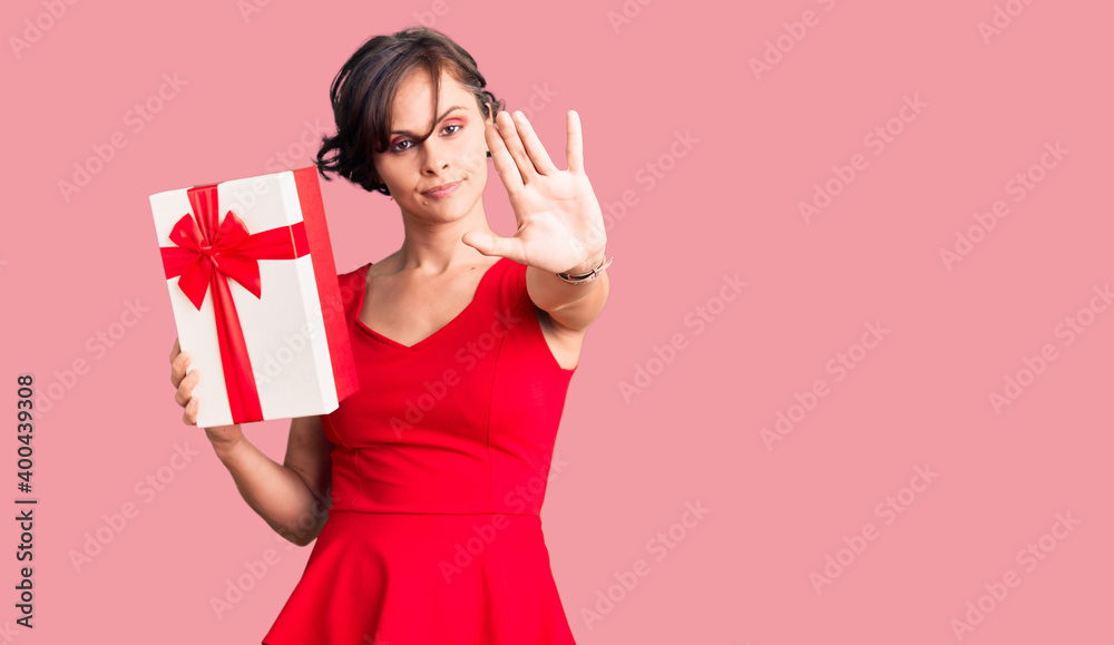 Beautiful young woman with short hair holding gift with open hand doing stop sign with serious and confident expression, defense gesture Beautiful young woman with short hair holding gift with open hand doing stop sign with serious and confident expression, defense gesture