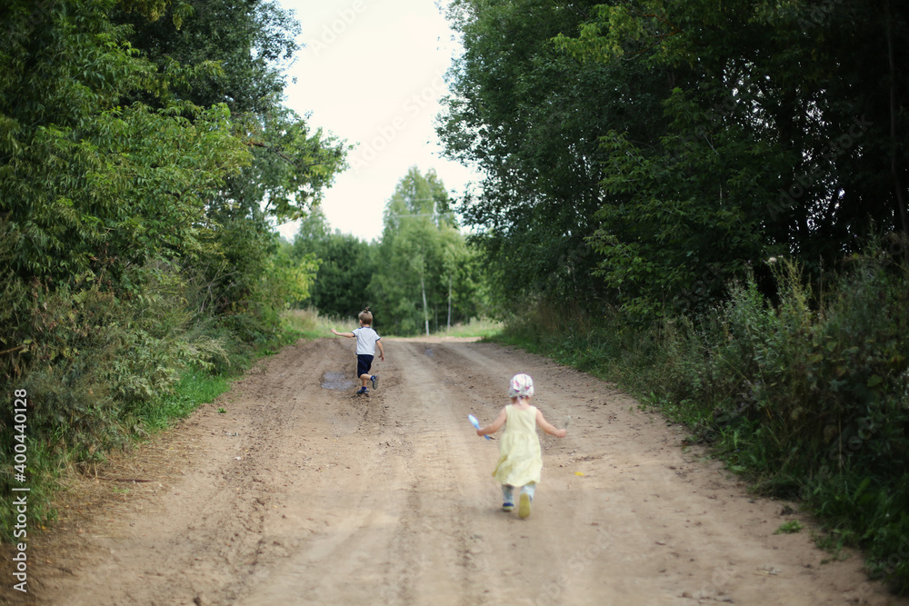 Sister in a yellow dress and brother are running on empty village road surrounded by the forest on a summer day. Siblings at summer vacations. Freedom in nature. Social distancing during coronavirus. Sister in a yellow dress and brother are running on empty village road surrounded by the forest on a summer day. Siblings at summer vacations. Freedom in nature. Social distancing during coronavirus.