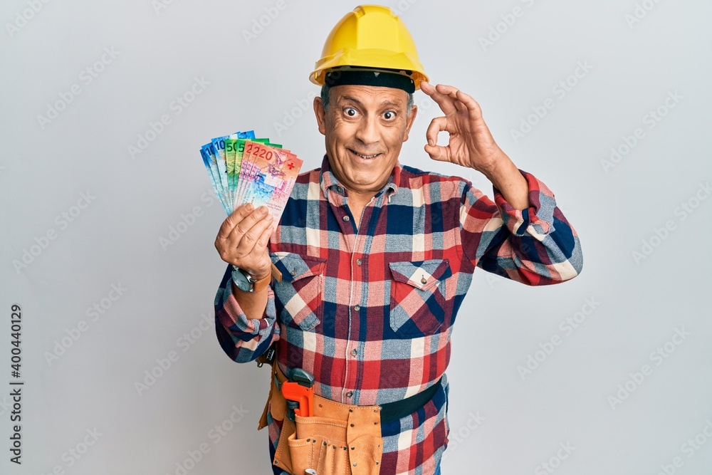 Senior hispanic man wearing handyman uniform and hardhat doing ok sign with fingers, smiling friendly gesturing excellent symbol