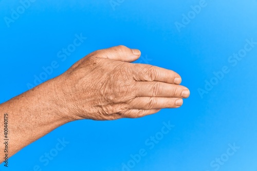 Hand of senior hispanic man over blue isolated background stretching and reaching with open hand for handshake, showing back of the hand © Krakenimages.com