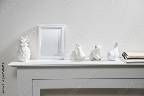 Figurines of a polar bear  pineapple  pears  an angel  a bullfinch bird and a white photo frame with empty space on a white fireplace console.