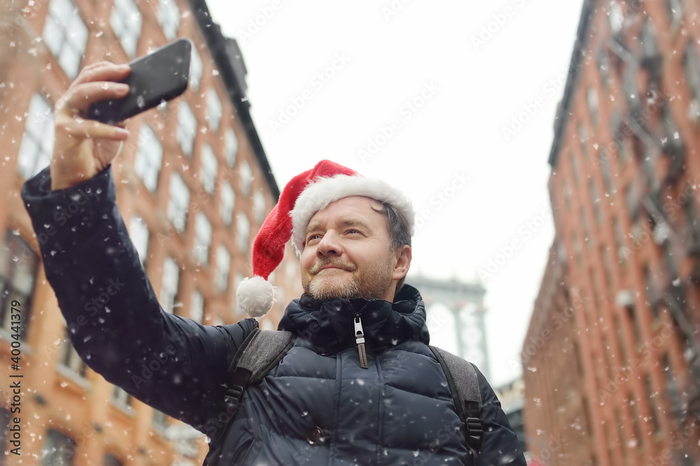 Obraz premium Middle aged man tourist in Santa Claus hat takes selfie on street near the Manhattan Bridge in New York City on Christmas Eve. Winter holidays in NYC.