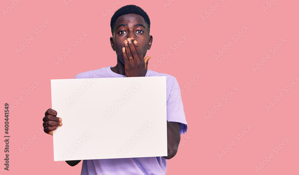 Young african american man holding blank empty banner covering mouth with hand, shocked and afraid for mistake. surprised expression