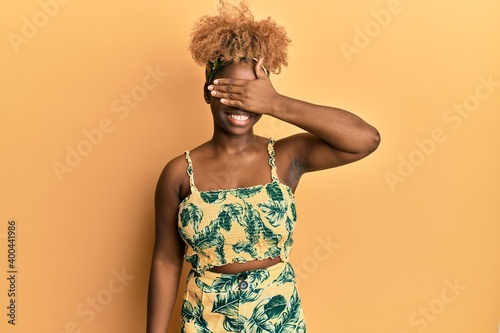 Young african woman with afro hair wearing summer dress smiling and laughing with hand on face covering eyes for surprise. blind concept.