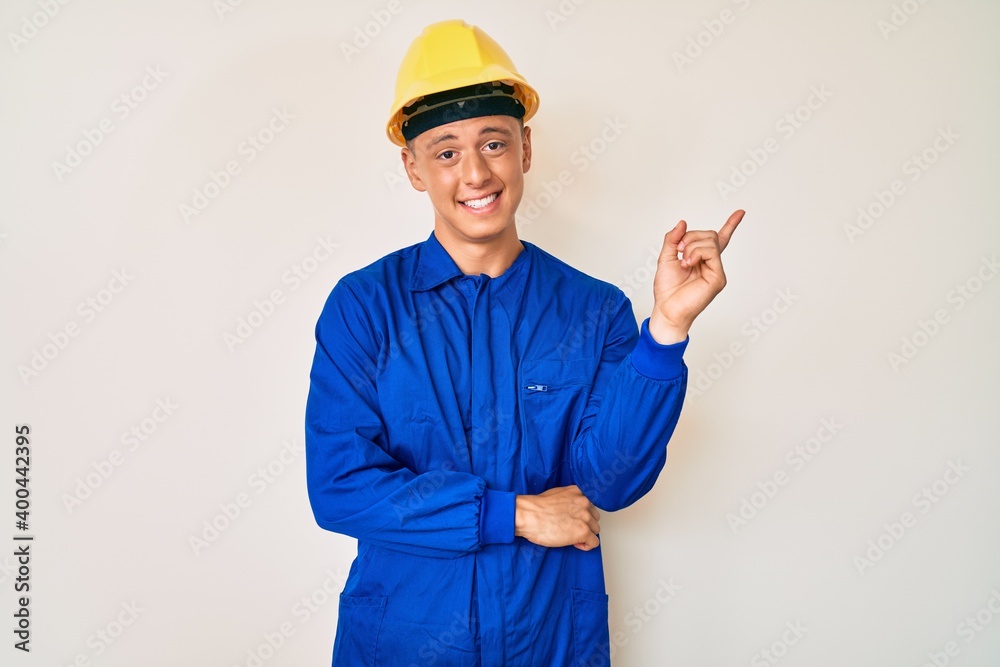 Young hispanic boy wearing worker uniform and hardhat smiling happy pointing with hand and finger to the side