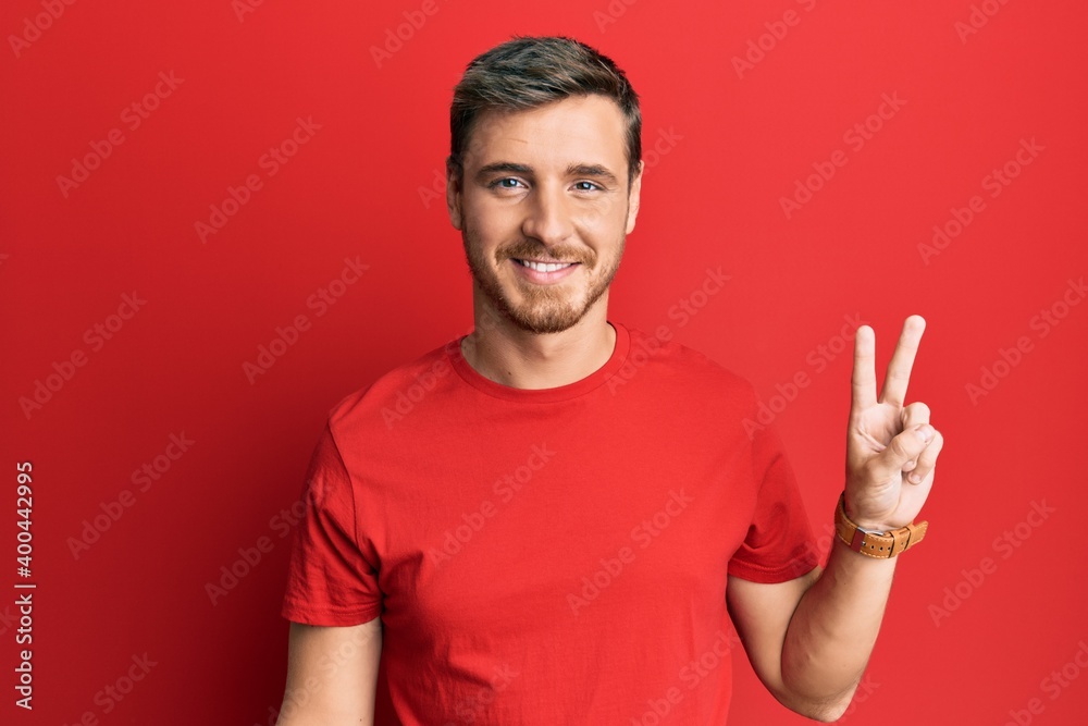 Handsome caucasian man wearing casual red tshirt smiling looking to the camera showing fingers doing victory sign. number two. Handsome caucasian man wearing casual red tshirt smiling looking to the camera showing fingers doing victory sign. number two.