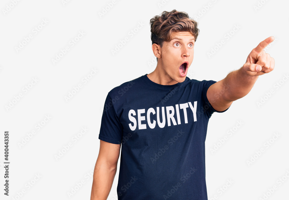 Young handsome man wearing security t shirt pointing with finger surprised ahead, open mouth amazed expression, something on the front Young handsome man wearing security t shirt pointing with finger surprised ahead, open mouth amazed expression, something on the front