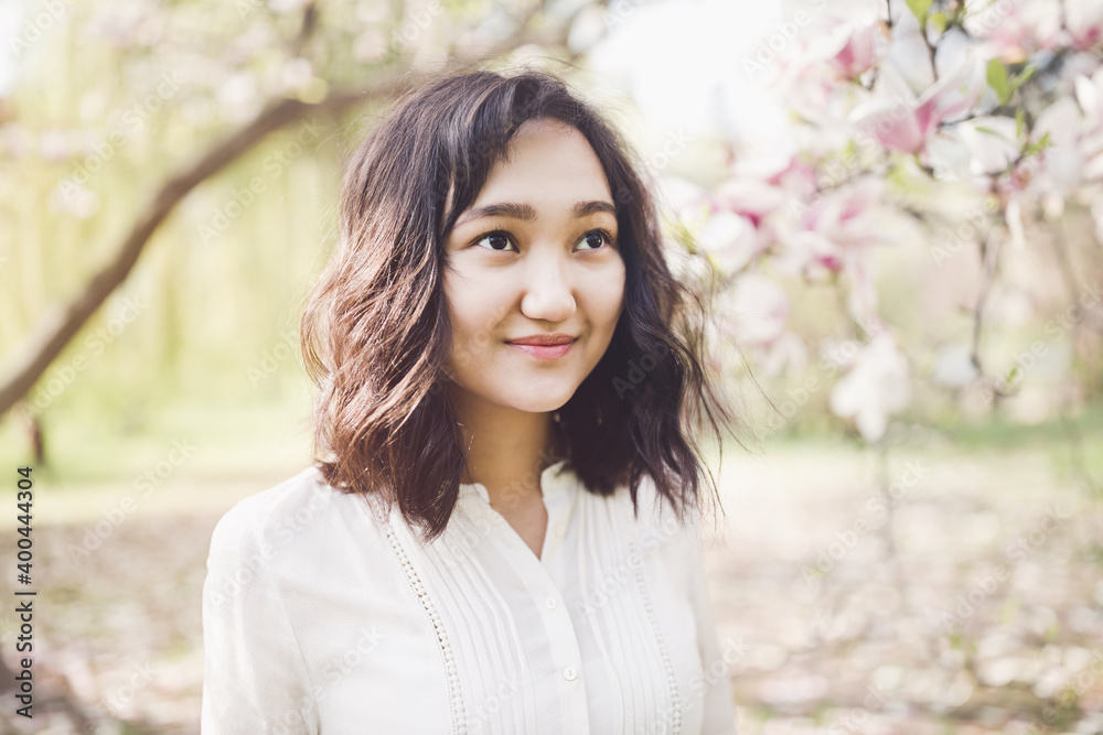 Young attractive asian woman in a white shirt under the blooming magnolia tree in park, calm, smiling. Spring portrait, lifestyle concept. Sunlight, copy space Young attractive asian woman in a white shirt under the blooming magnolia tree in park, calm, smiling. Spring portrait, lifestyle concept. Sunlight, copy space