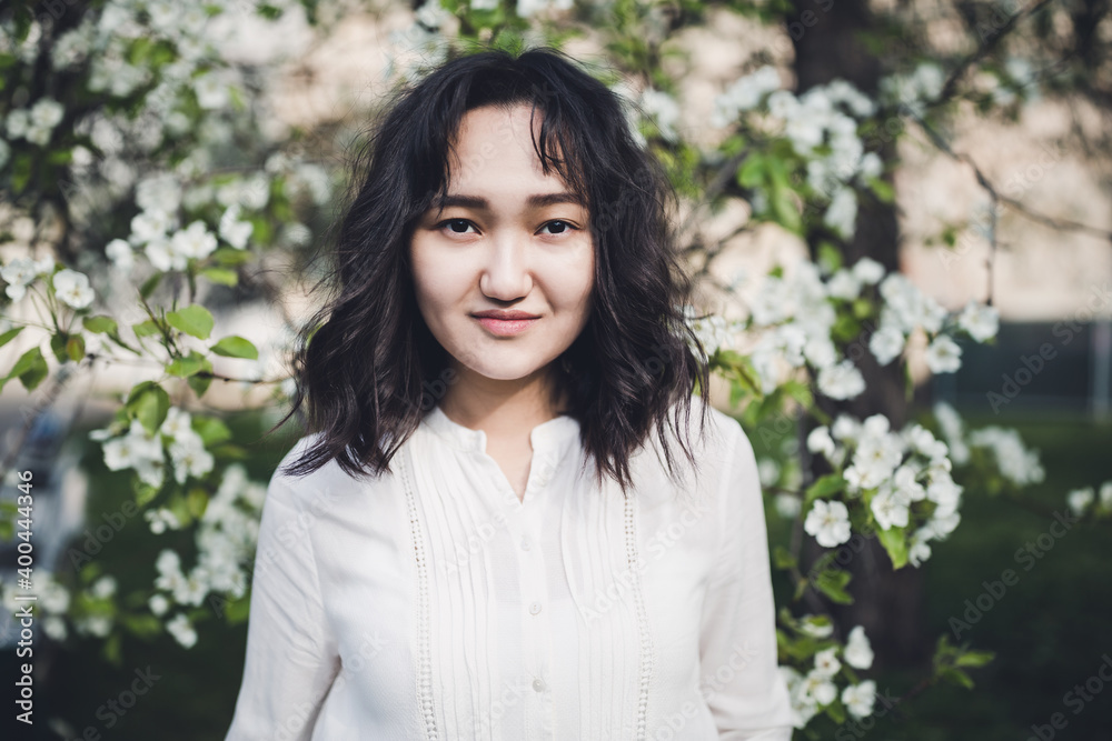 Young attractive asian woman in a white shirt under the blooming tree in park, calm, smiling. Spring portrait, lifestyle concept. Moody picture, copy space Young attractive asian woman in a white shirt under the blooming tree in park, calm, smiling. Spring portrait, lifestyle concept. Moody picture, copy space