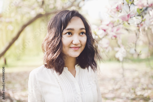 Young attractive asian woman in a white shirt under the blooming magnolia tree in park, calm, smiling. Spring portrait, lifestyle concept. Sunlight, copy space photo