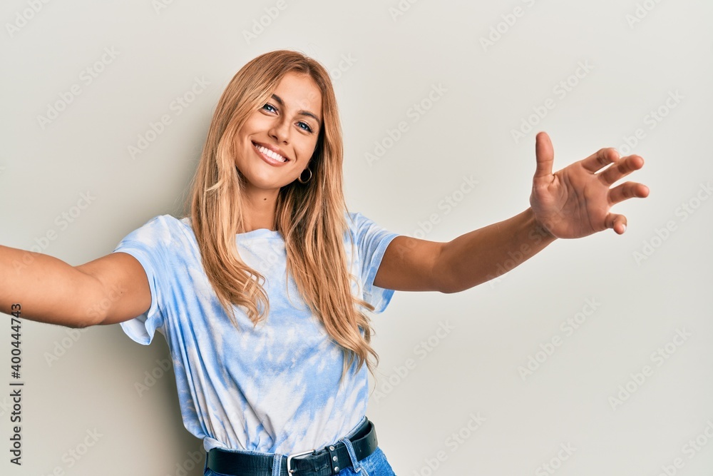 Beautiful blonde young woman wearing tye die tshirt looking at the camera smiling with open arms for hug. cheerful expression embracing happiness. Beautiful blonde young woman wearing tye die tshirt looking at the camera smiling with open arms for hug. cheerful expression embracing happiness.