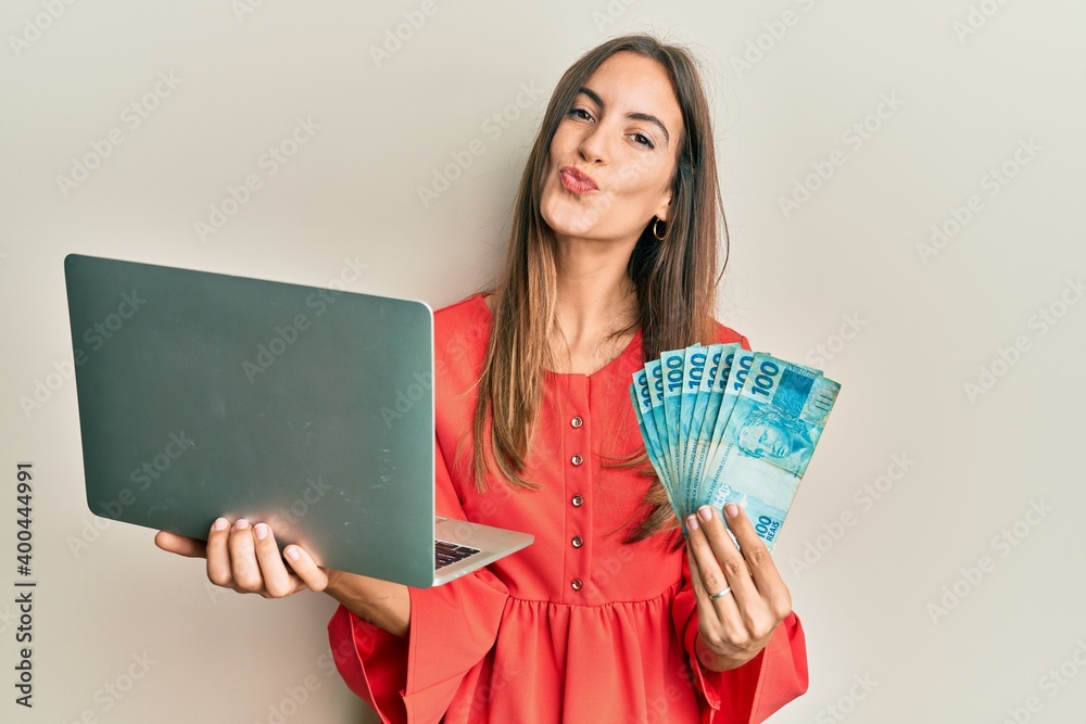 Young beautiful woman holding laptop and holding brazilian real looking at the camera blowing a kiss being lovely and sexy. love expression. Young beautiful woman holding laptop and holding brazilian real looking at the camera blowing a kiss being lovely and sexy. love expression.