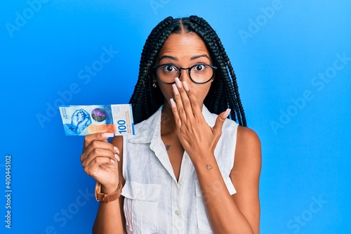 Beautiful hispanic woman holding 100 swiss franc banknote covering mouth with hand, shocked and afraid for mistake. surprised expression